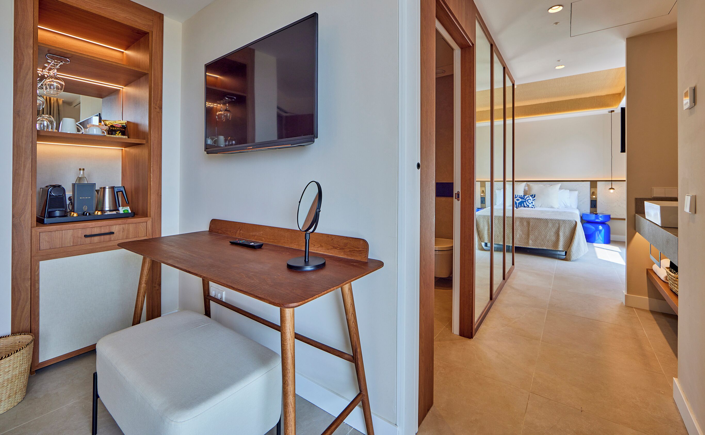 Habitación de hotel con un escritorio de madera, un televisor, una cama con detalles azules y un baño visible a través de una puerta de cristal.