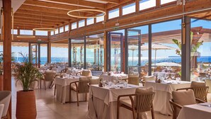 A restaurant with multiple tables and chairs, large windows offering a view of the sea, and a wooden ceiling. - Universal Grand León & Spa (Ses Salines)