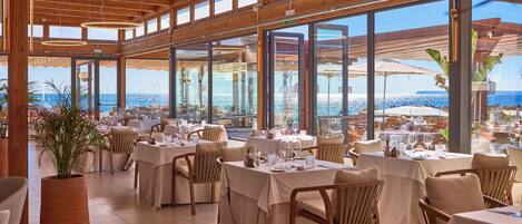 Ein Restaurant mit mehreren Tischen und Stühlen, großen Fenstern mit Meerblick und einer Holzkονstruktion an der Decke.