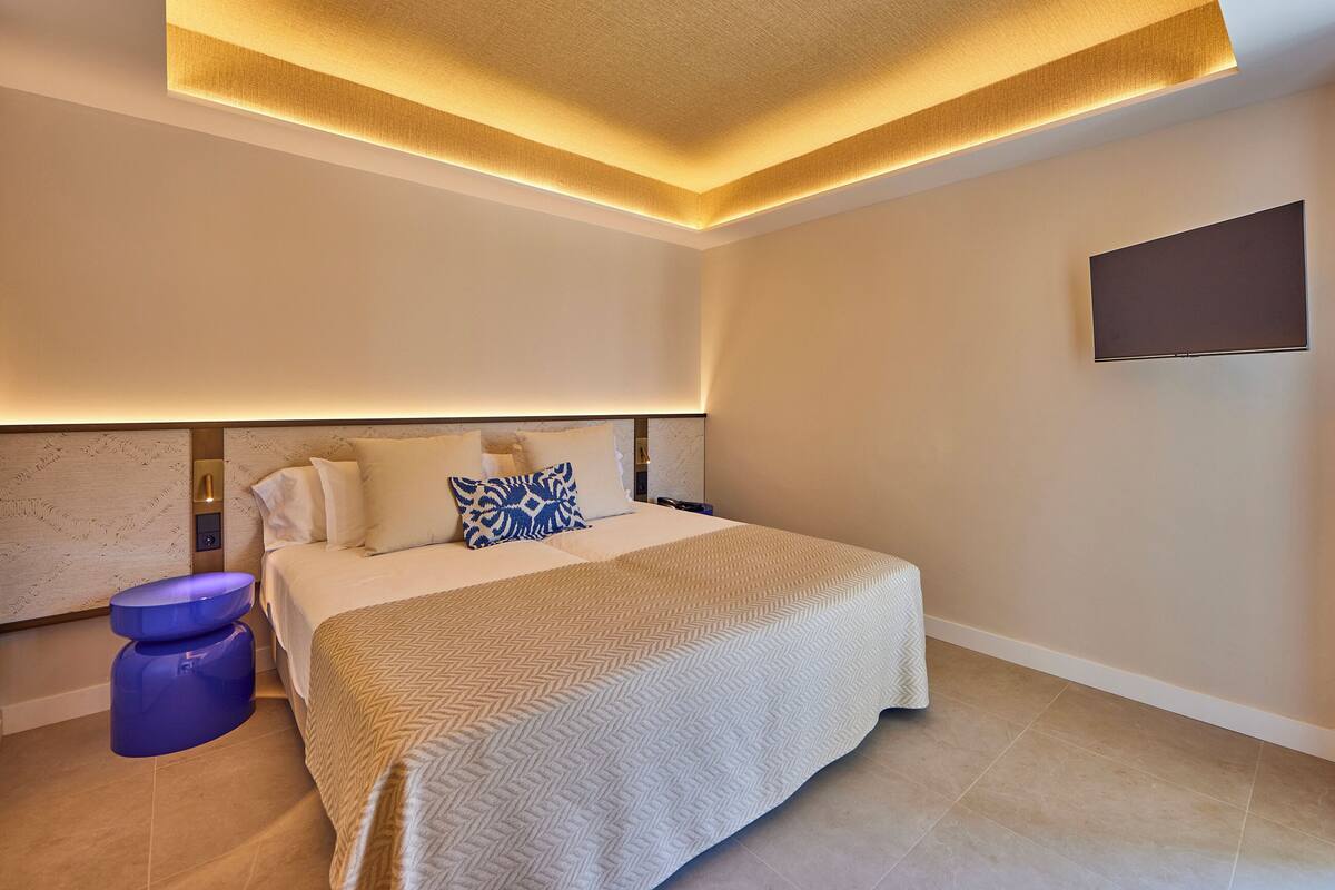 A modern hotel room with a large bed, a flat-screen TV mounted on the wall, and a unique ceiling design.