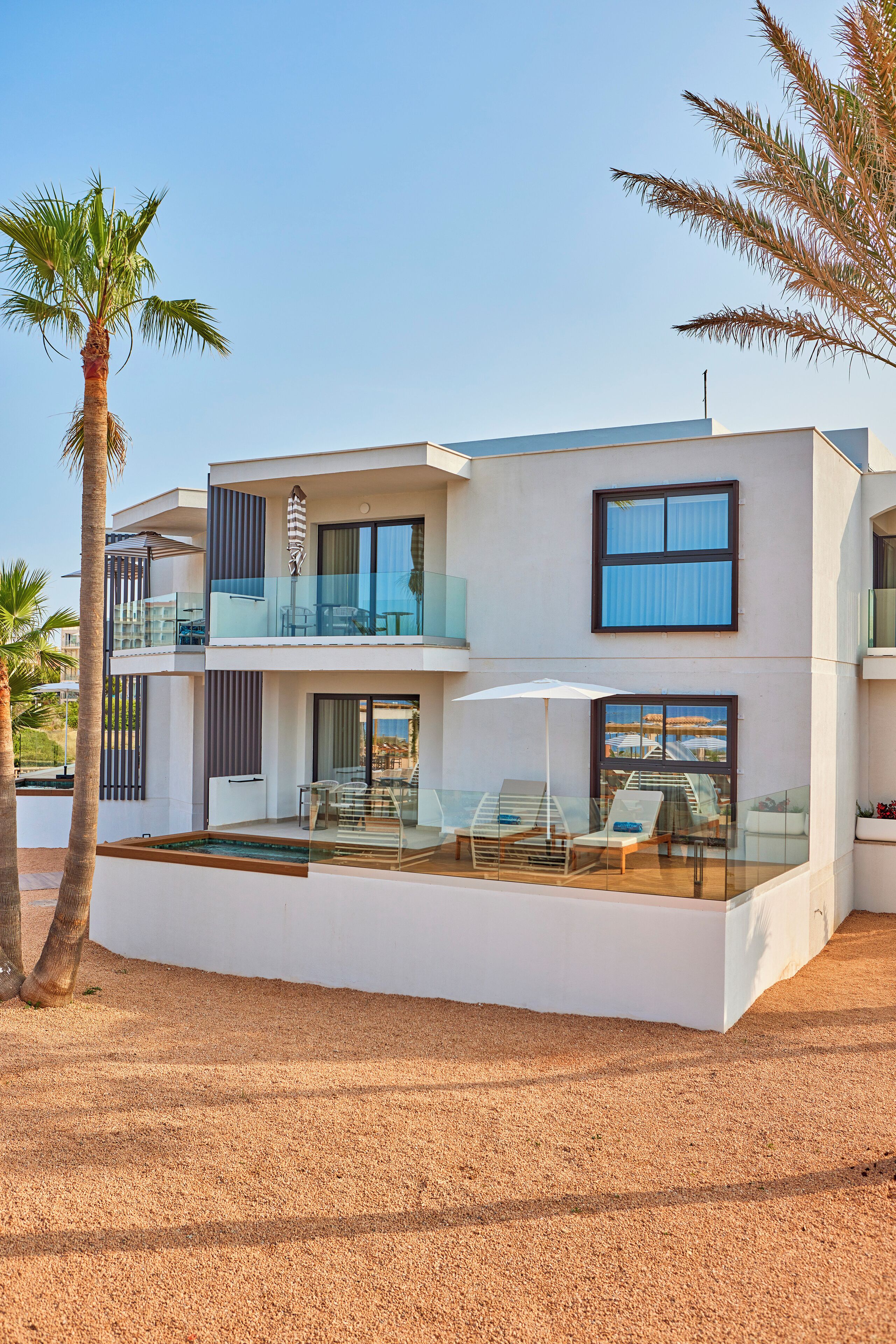 A modern two-story house with a glass balcony, a pool area, and palm trees.