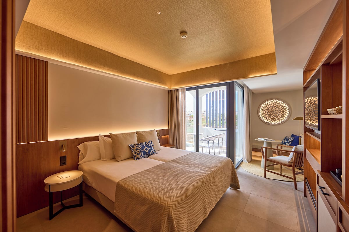 A modern hotel room with a large bed, a small side table, a seating area with a table and chairs, and a large window with a view.