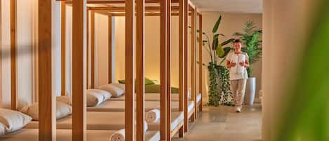 Habitación de hotel con camas de madera, una planta grande en maceta y una persona caminando por el pasillo.