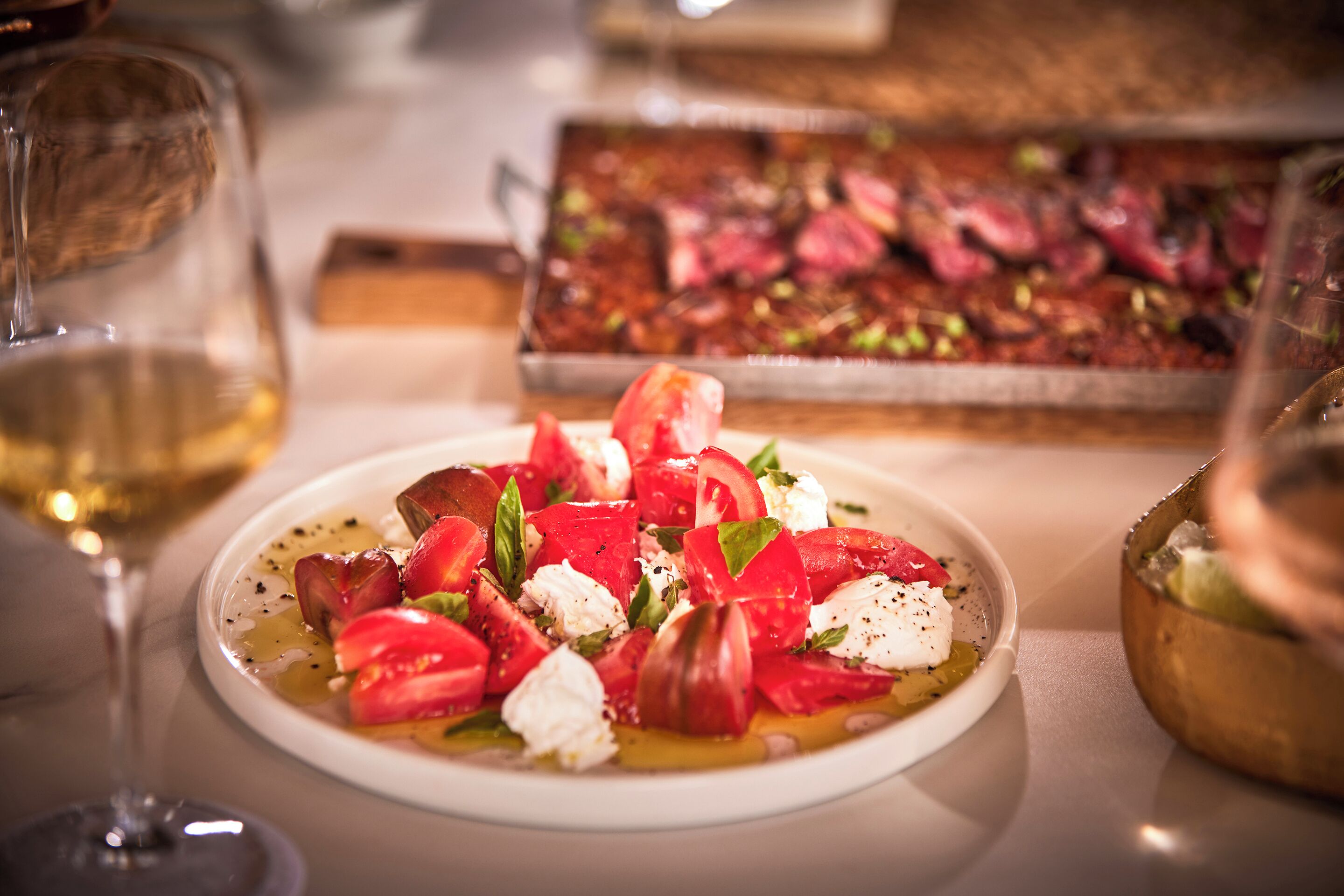 A plate of Caprese salad with tomatoes, mozzarella, and basil, a glass of white wine, and a roasted meat dish in the background.