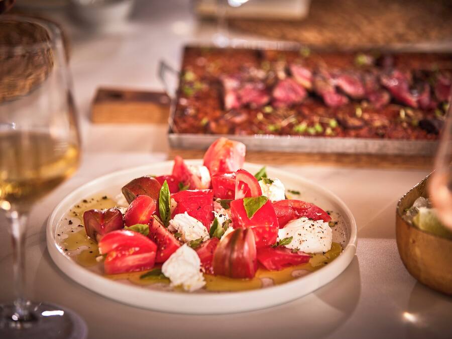 A plate of Caprese salad with tomatoes, mozzarella, and basil, a glass of white wine, and a roasted meat dish in the background.