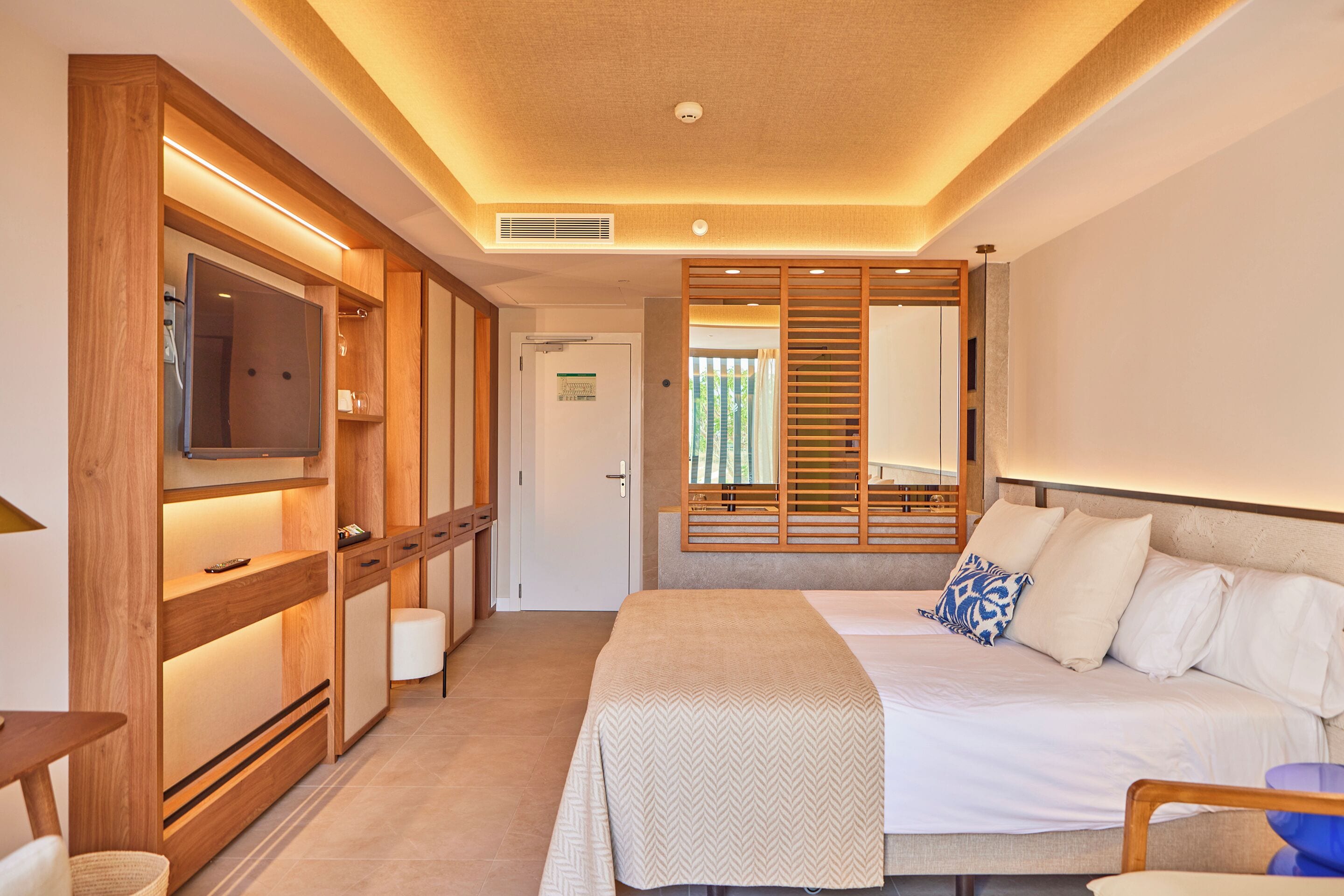 Una habitación de hotel moderna con una cama grande, un televisor, muebles de madera y un ventanal corredero que da a un balcón.