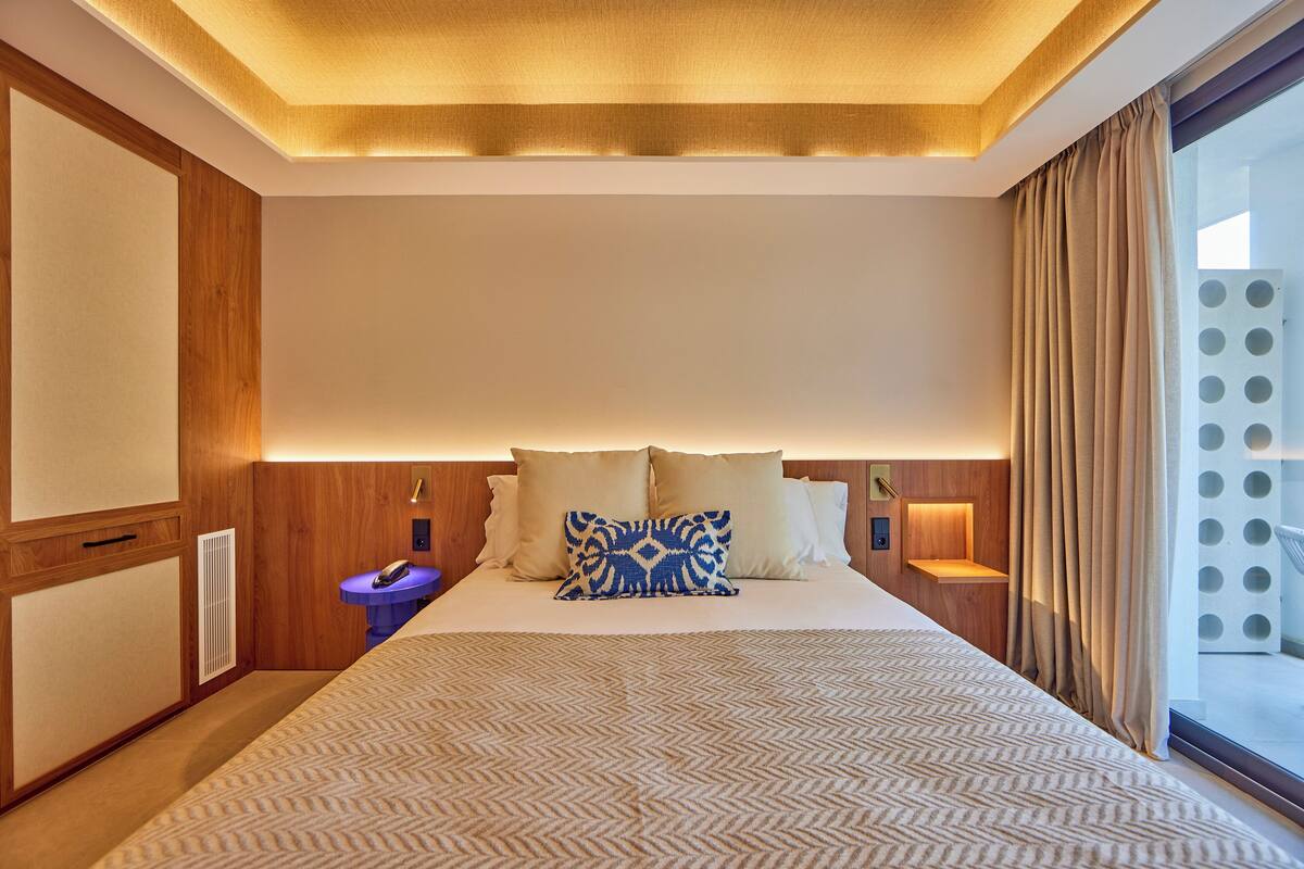A hotel room with a large bed, wooden paneling, a bedside table with a lamp, and a view of the outside through a large window.