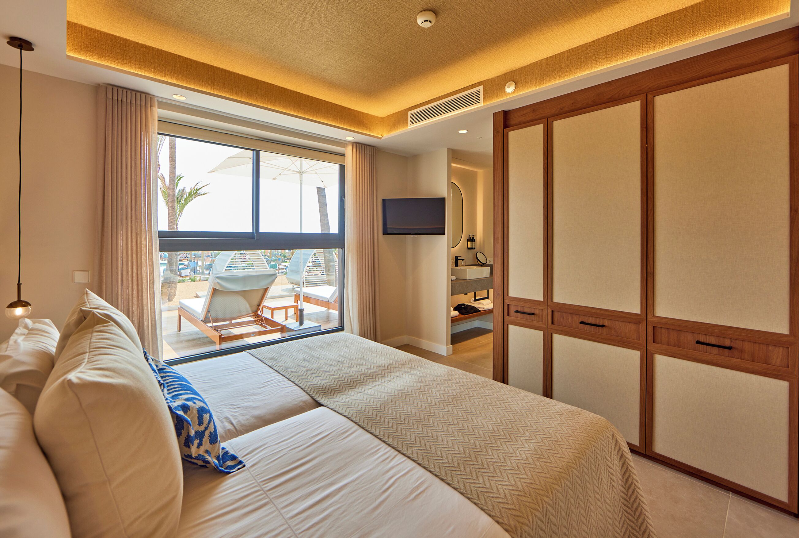 A hotel room with a large bed, a TV, a sliding door closet, and a balcony with a view of palm trees.
