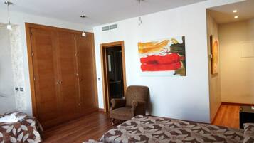A bedroom with a wooden wardrobe, a chair, a bed with a leaf-patterned bedspread, and a painting on the wall.