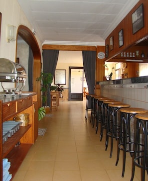 Un restaurante alargado y estrecho con mesas y sillas de madera, una estación de buffet y una zona de cocina.