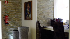 A room with a stone wall, a fire extinguisher, a framed painting, a desk with chairs, and a window with books on the sill. - Guest House Bocage (Setubal)