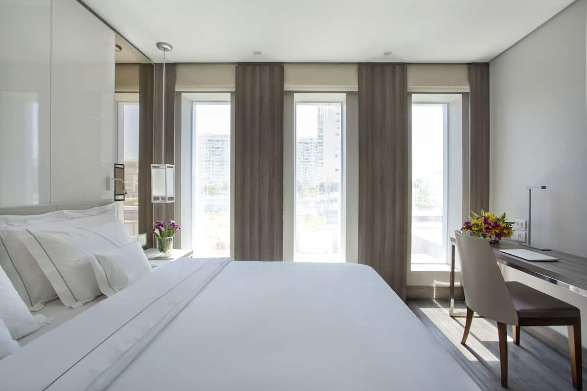 A modern hotel room with a large bed, a desk, and a chair. There are curtains on the windows, and a vase with flowers on the desk.