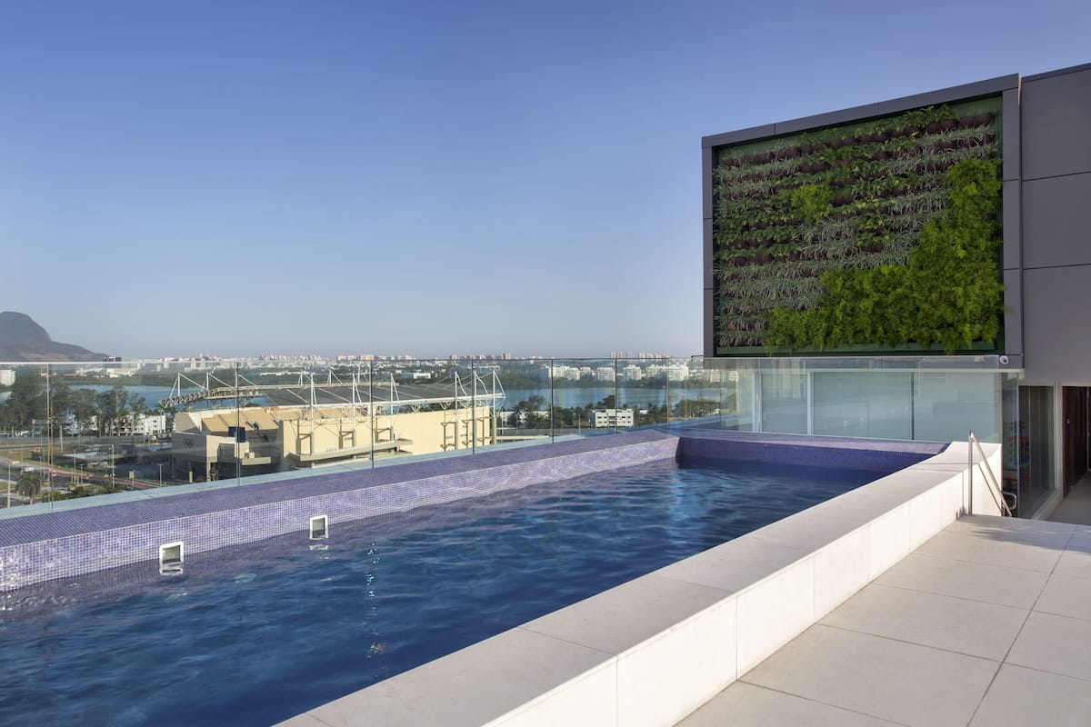 A rooftop infinity pool with a city view, a glass wall with greenery, and a clear sky.
