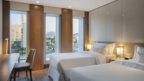 Quarto de hotel com duas camas, uma escrivaninha e uma cadeira. Janelas amplas com vista para a cidade.
