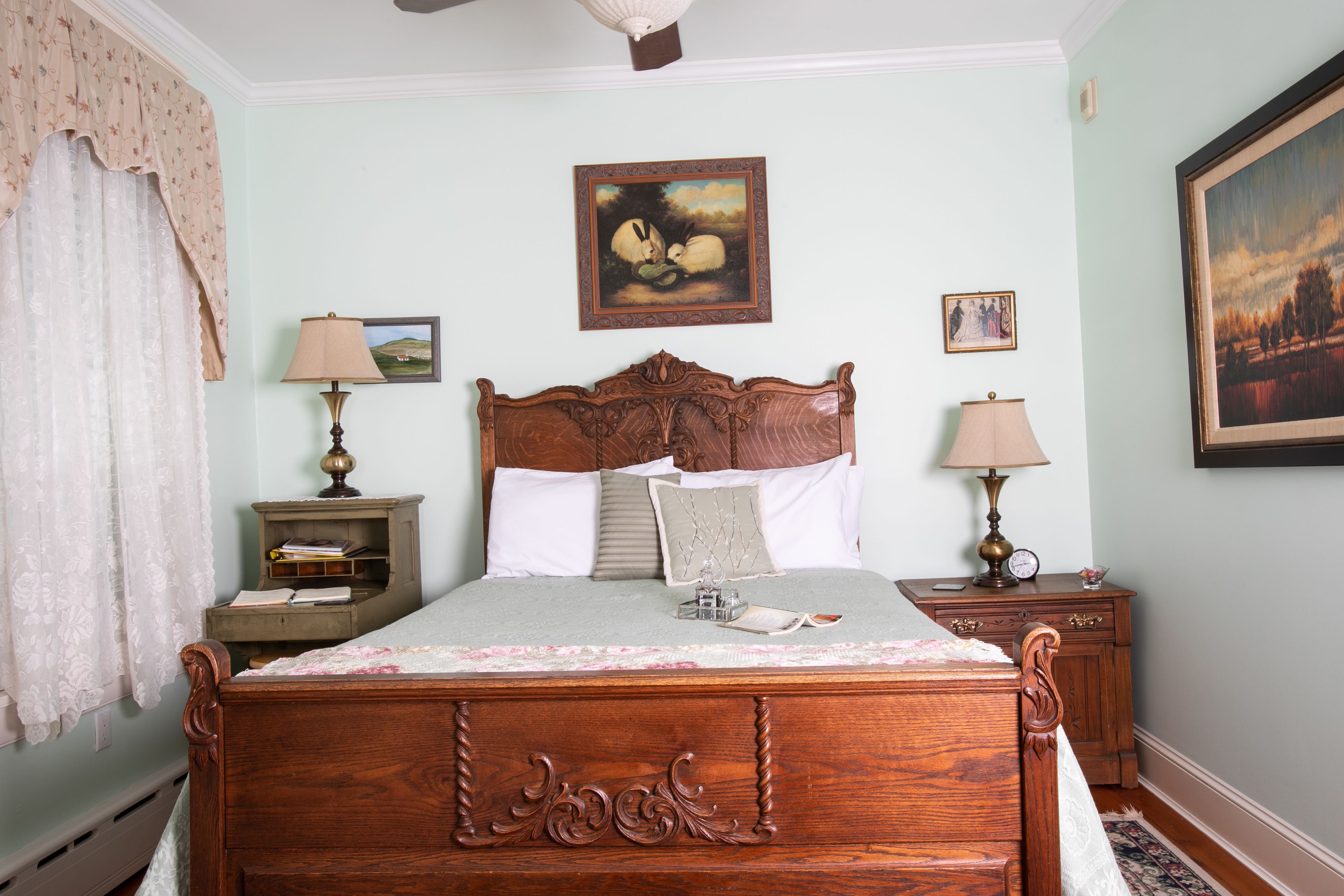 A traditional bedroom with a wooden bed, two bedside tables with lamps, a ceiling fan, and framed artwork on the walls.