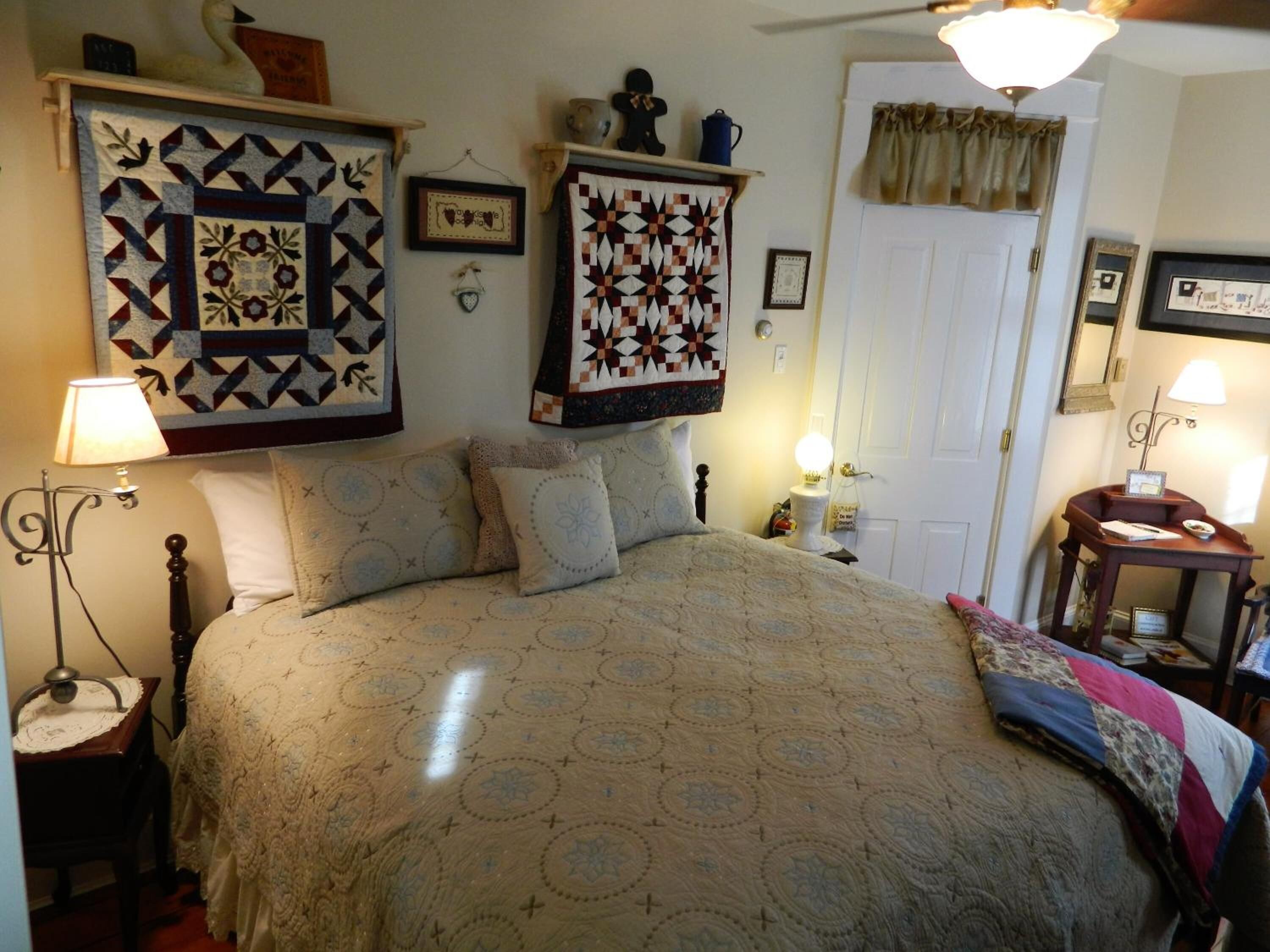 A bedroom with a large bed, a nightstand, a lamp, a dresser, and a quilted wall hanging.
