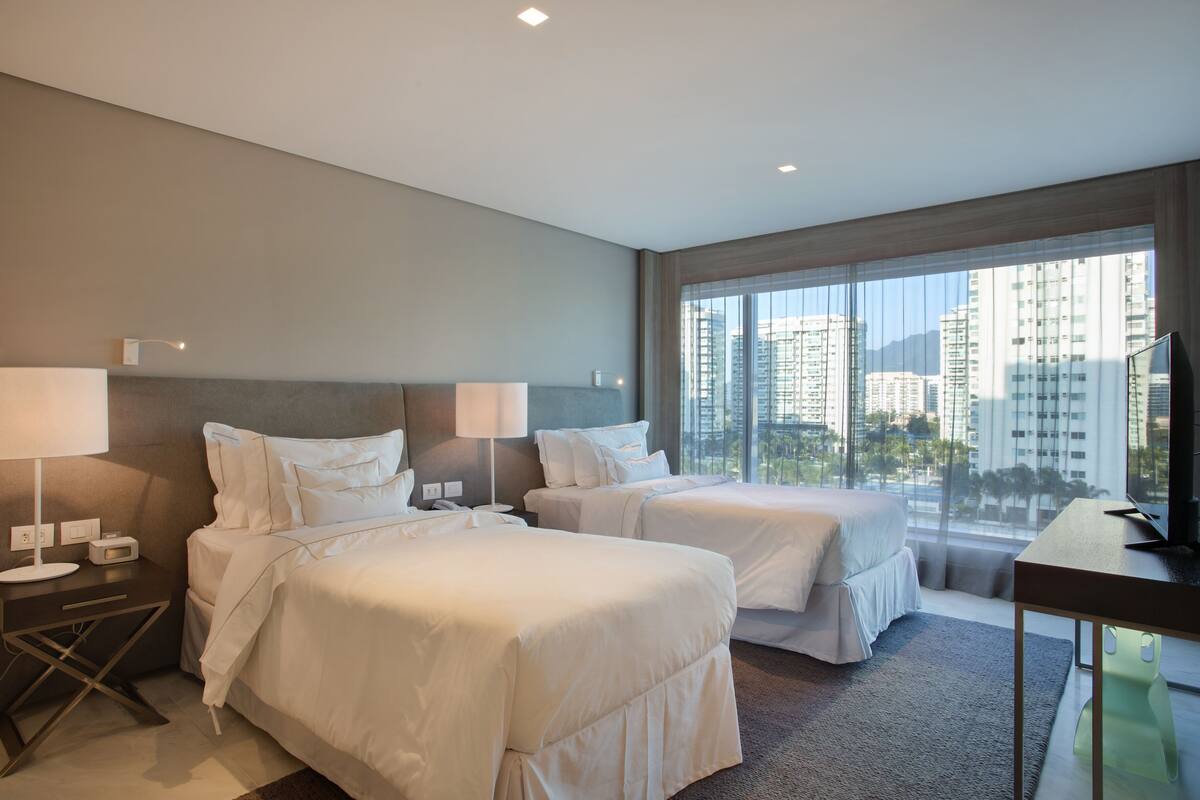Quarto de hotel com duas camas, uma escrivaninha, televisão e janelas amplas com vista para a cidade.