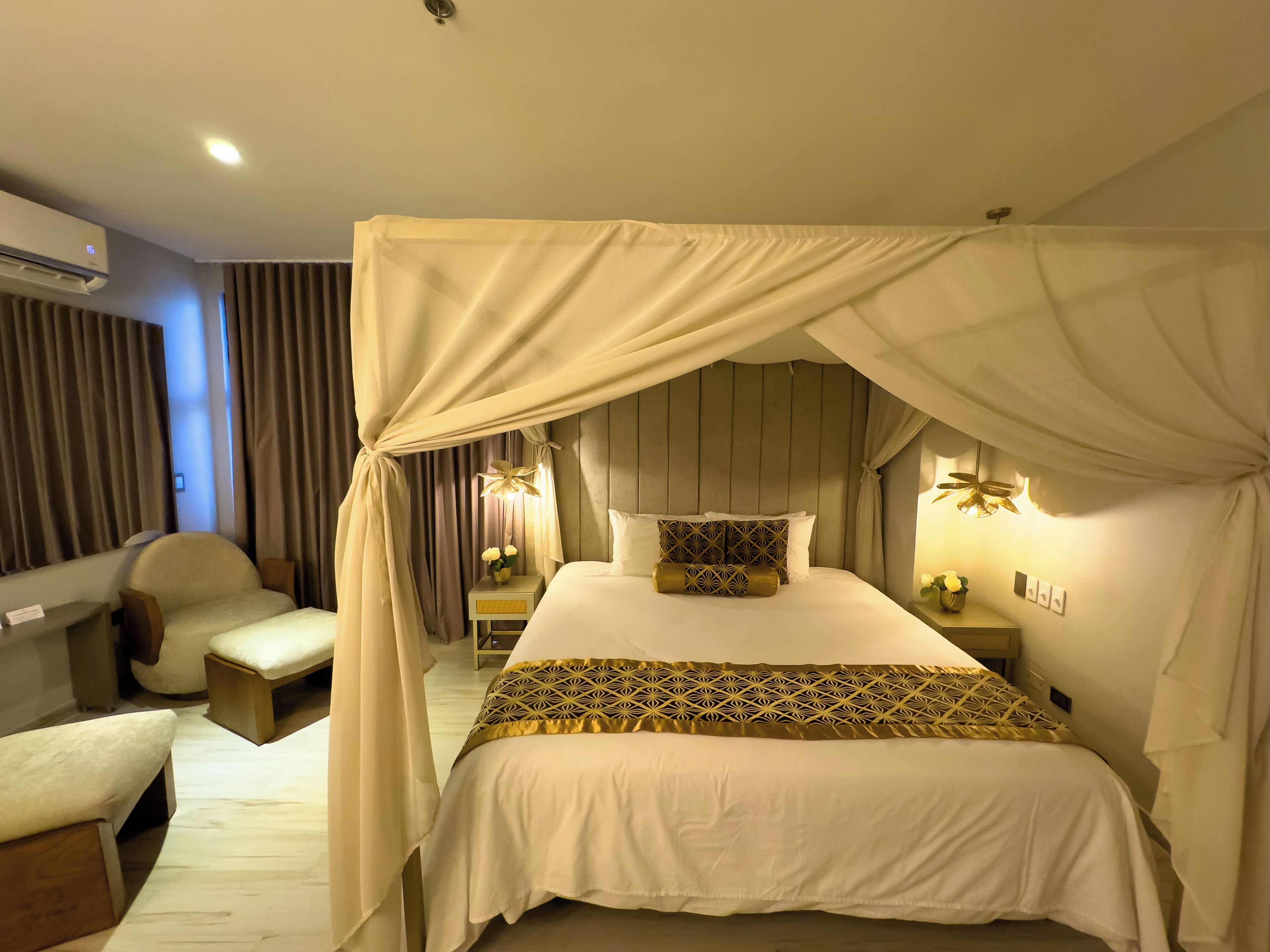 A bed with a canopy, bedside tables with lamps, a chair, and a wall-mounted air conditioner.