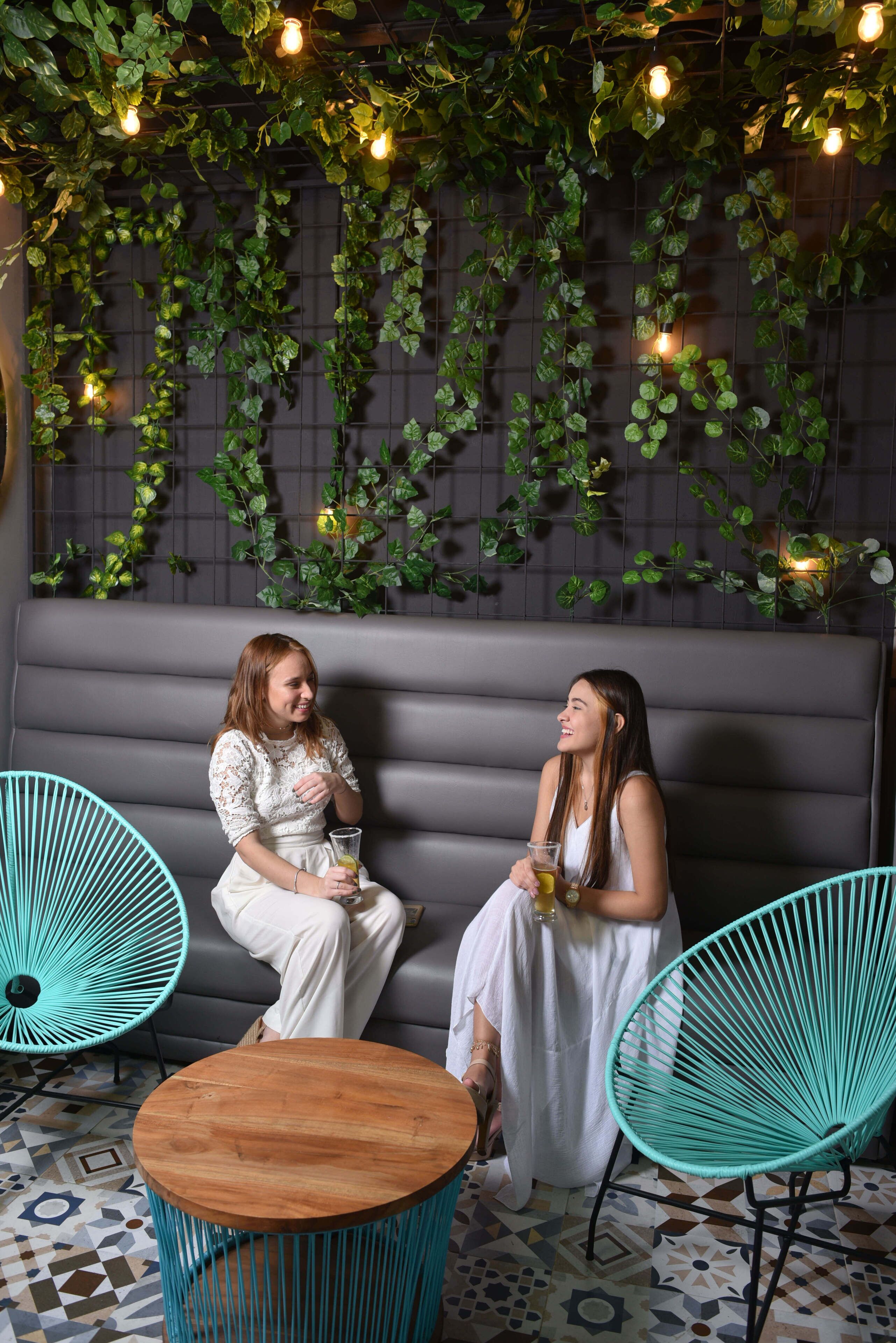 Dos personas sentadas en un sofá, con un suelo estampado y una pared decorada con enredaderas verdes y luces colgantes.