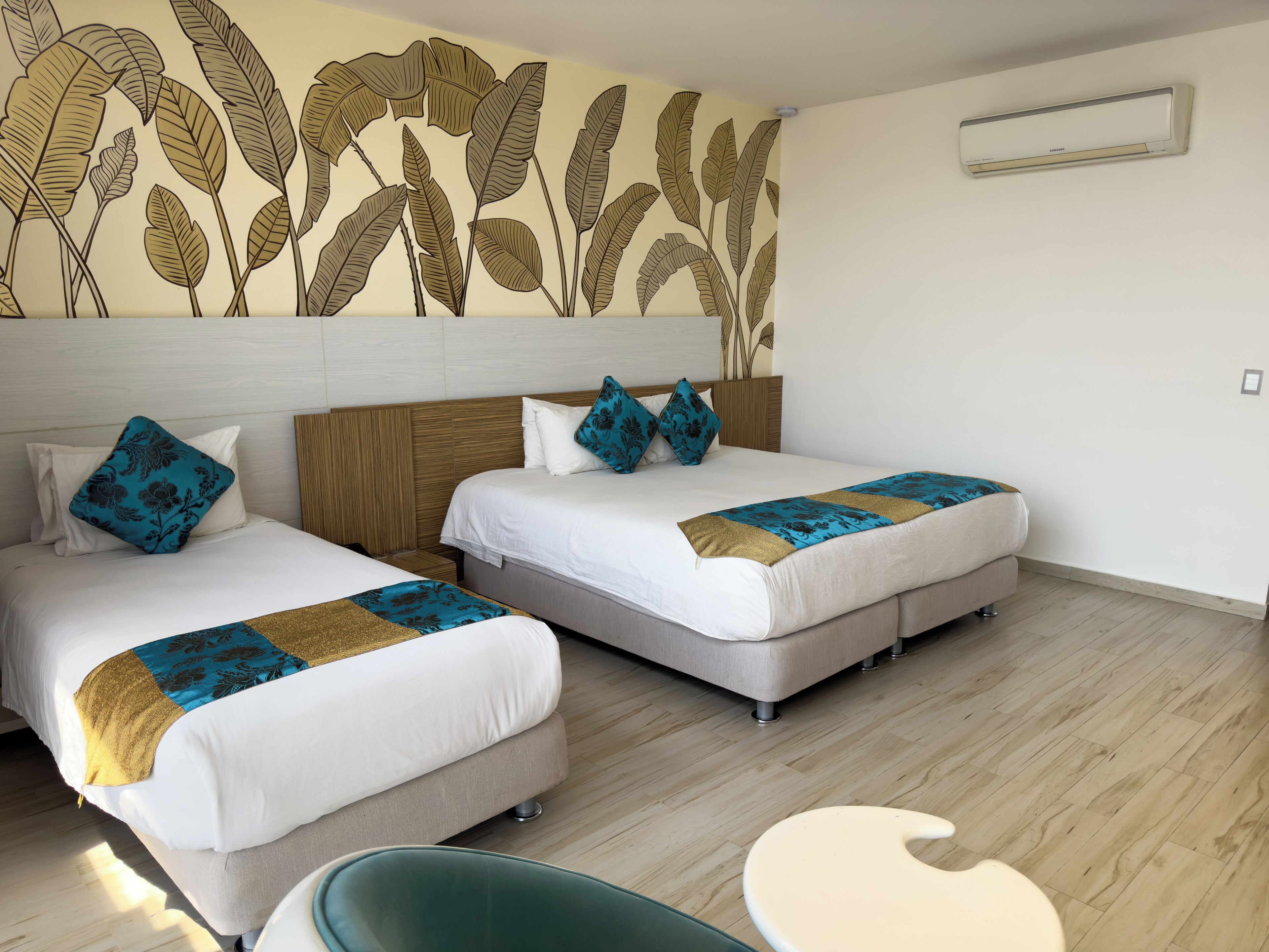Habitación de hotel con dos camas, cabecera de madera, un mural en la pared, un equipo de aire acondicionado y una pequeña mesa redonda.