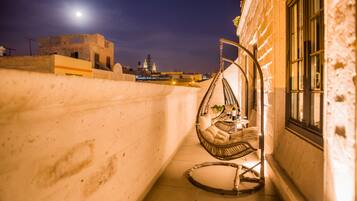 A balcony with a hanging chair, a small table with a plant, and a view of a building with a moon in the background.