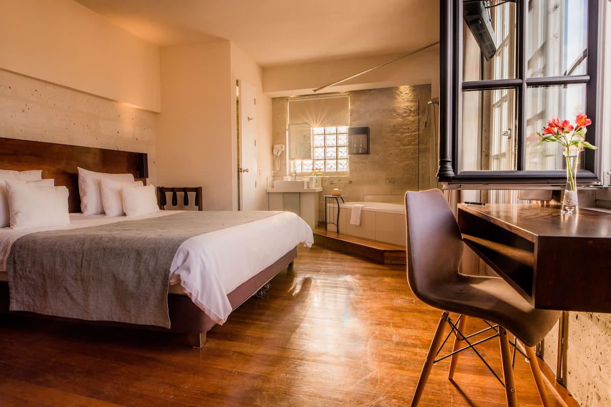 A bedroom with a large bed, a desk, and a chair. There is a window with a view of the outside.