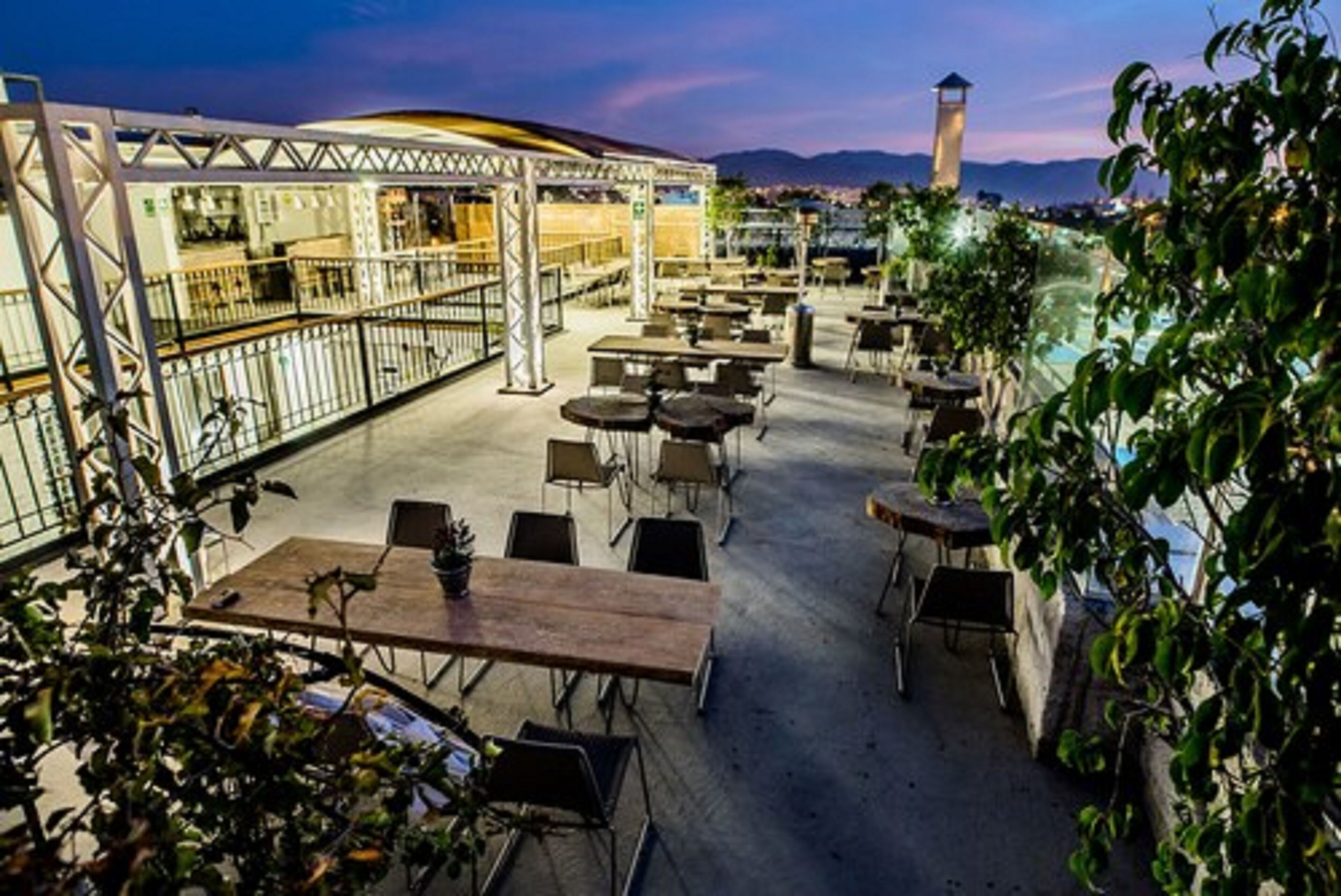 Un patio al aire libre con mesas y sillas, un área de estar cubierta y vista panorámica de la ciudad al fondo.