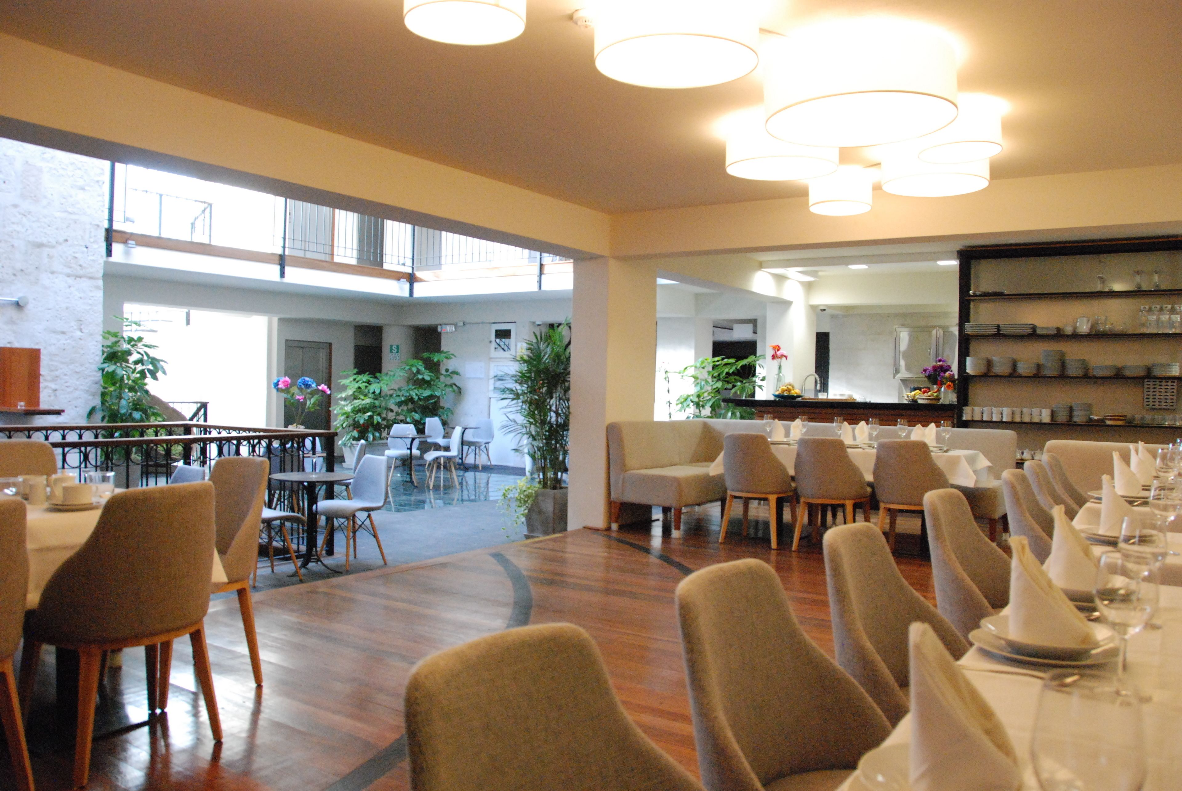 A spacious dining area with high ceilings and natural light, featuring multiple dining tables and chairs, a visible balcony, and a variety of plants.