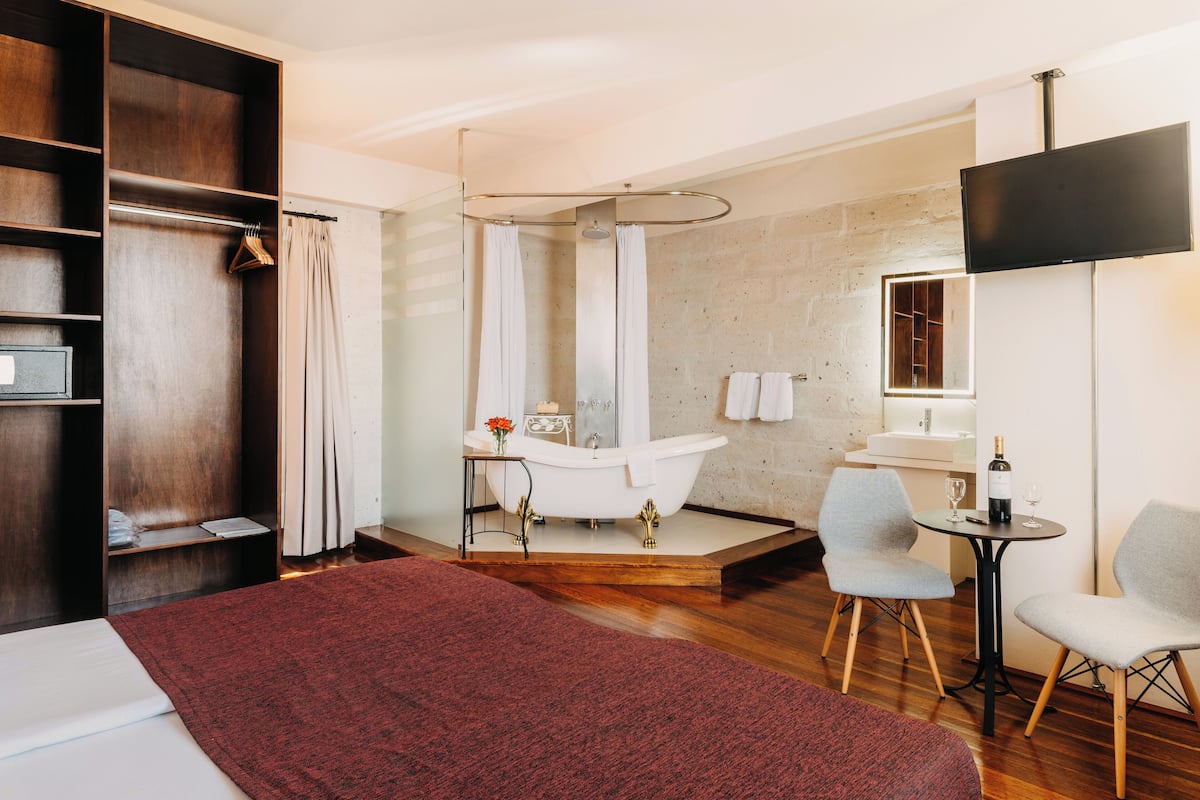 A modern bathroom with a freestanding bathtub, a shower, and a vanity with a mirror. There is a wooden floor, a red rug, and a small table with a bottle and glass.