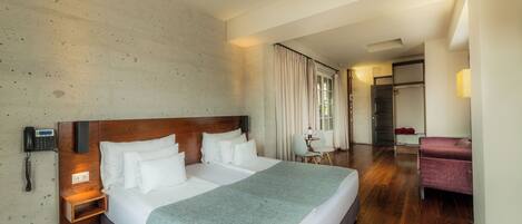 Habitación de hotel con una cama grande, un teléfono en la mesita de noche, una pequeña mesa con una silla y un sillón púrpura.