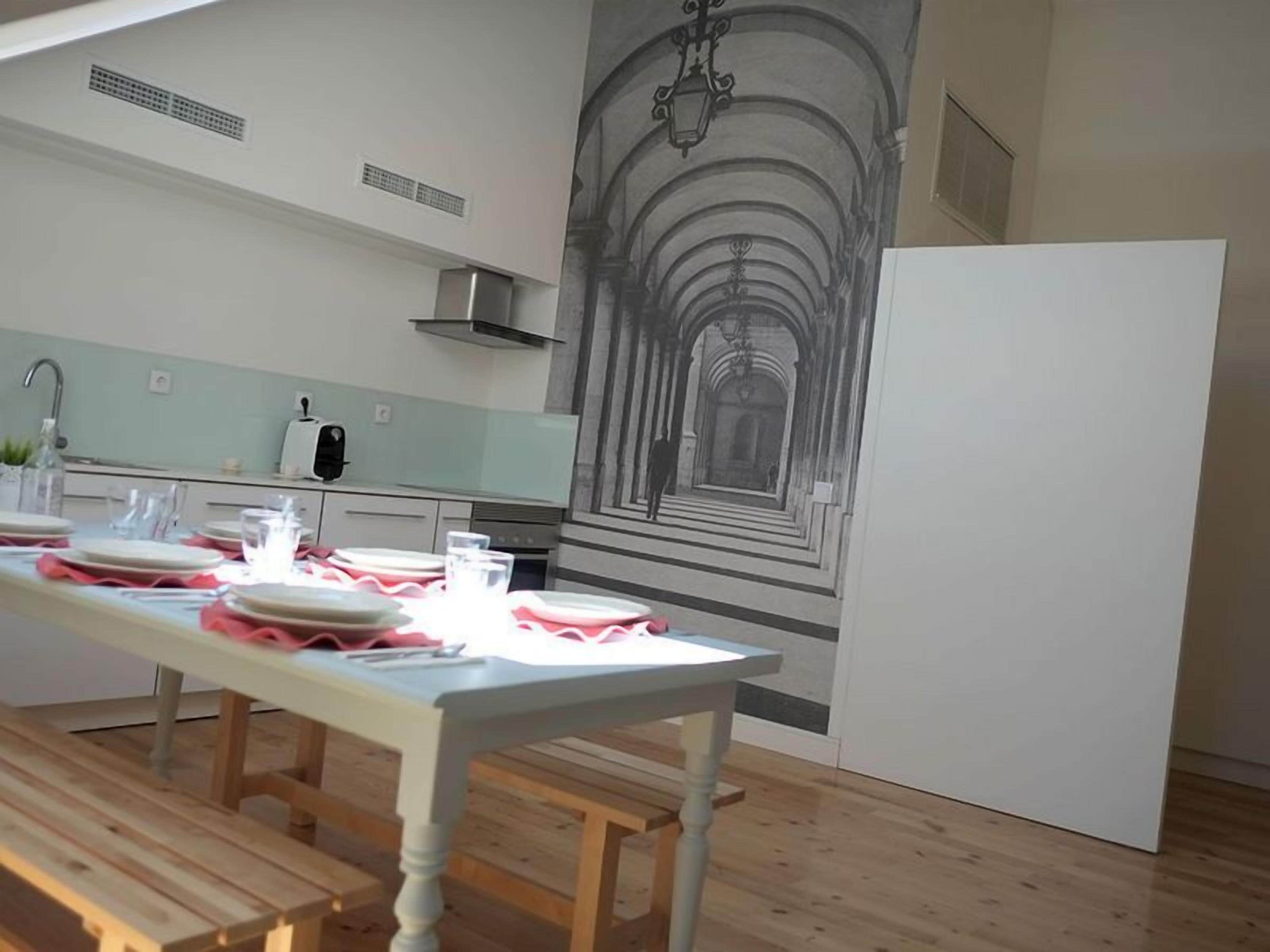A modern kitchen with a long dining table set for four, a white refrigerator, and a mural of a spiral staircase.