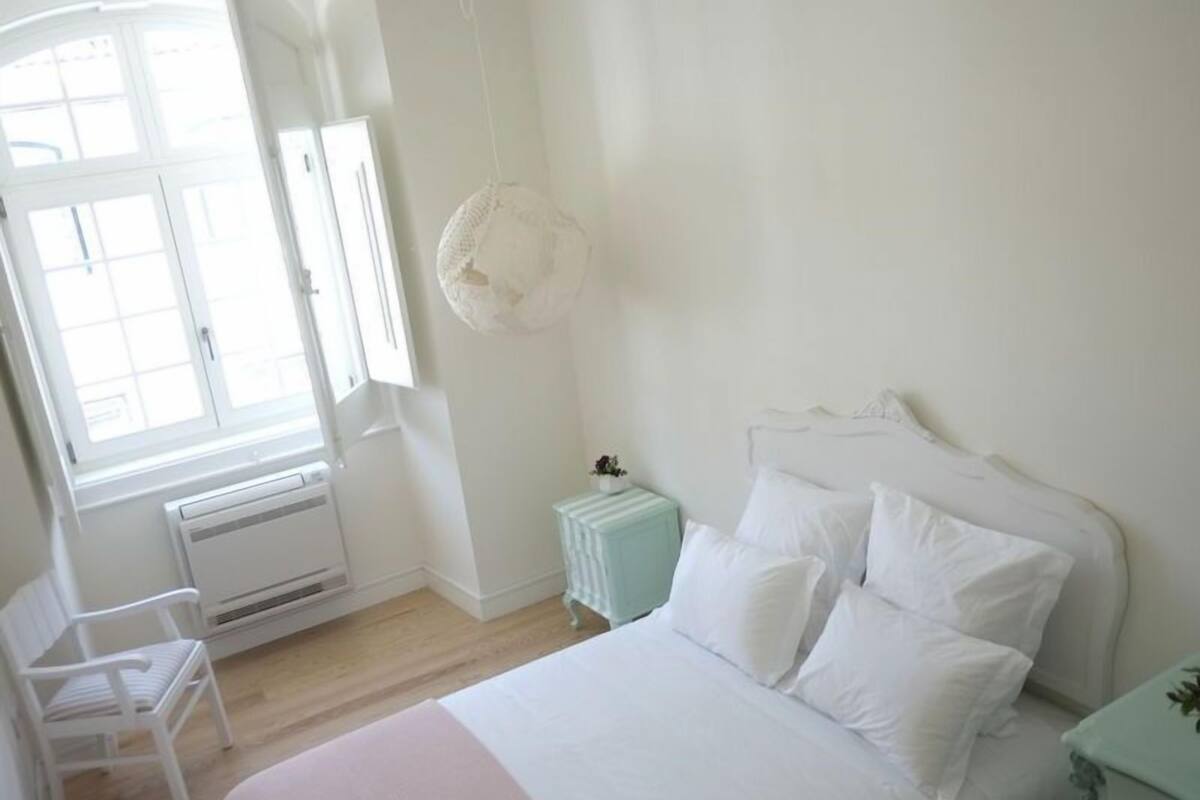 Ein Schlafzimmer mit Bett, Stuhl, kleinem Tisch, Pendelleuchte und Fenster.