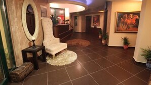A lobby with a tufted armchair, a round mirror, a clock, and a painting of horses. - Delfini Hotel (Amfilochia)