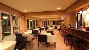 A restaurant with multiple tables and chairs, a bar area with stools, and large windows providing views of the outdoors. - Delfini Hotel (Amfilochia)