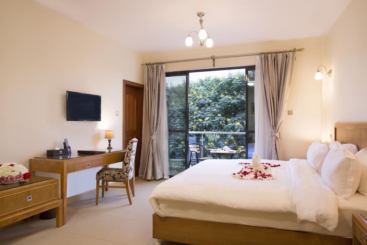 A hotel room with a bed, desk, chair, and a balcony with a view of trees.