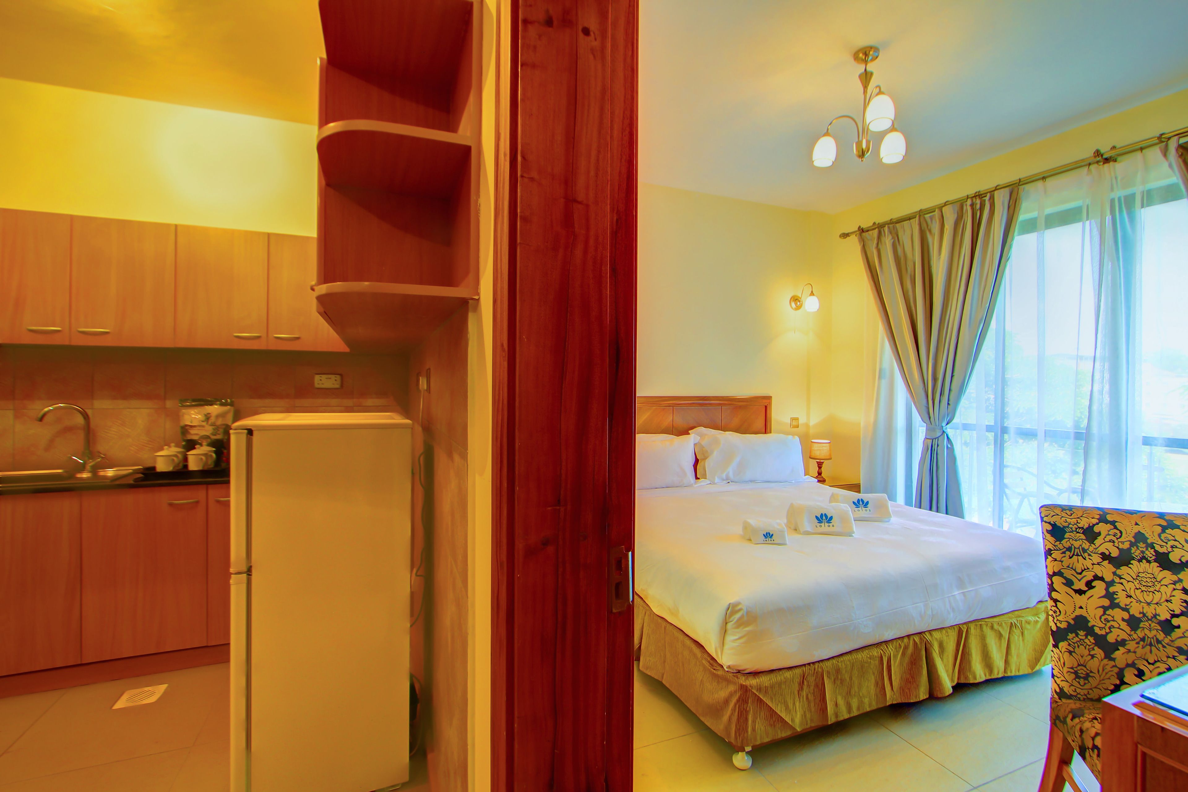 A hotel room with a bed, a desk, a chair, a kitchen area with a refrigerator, and a window with curtains.