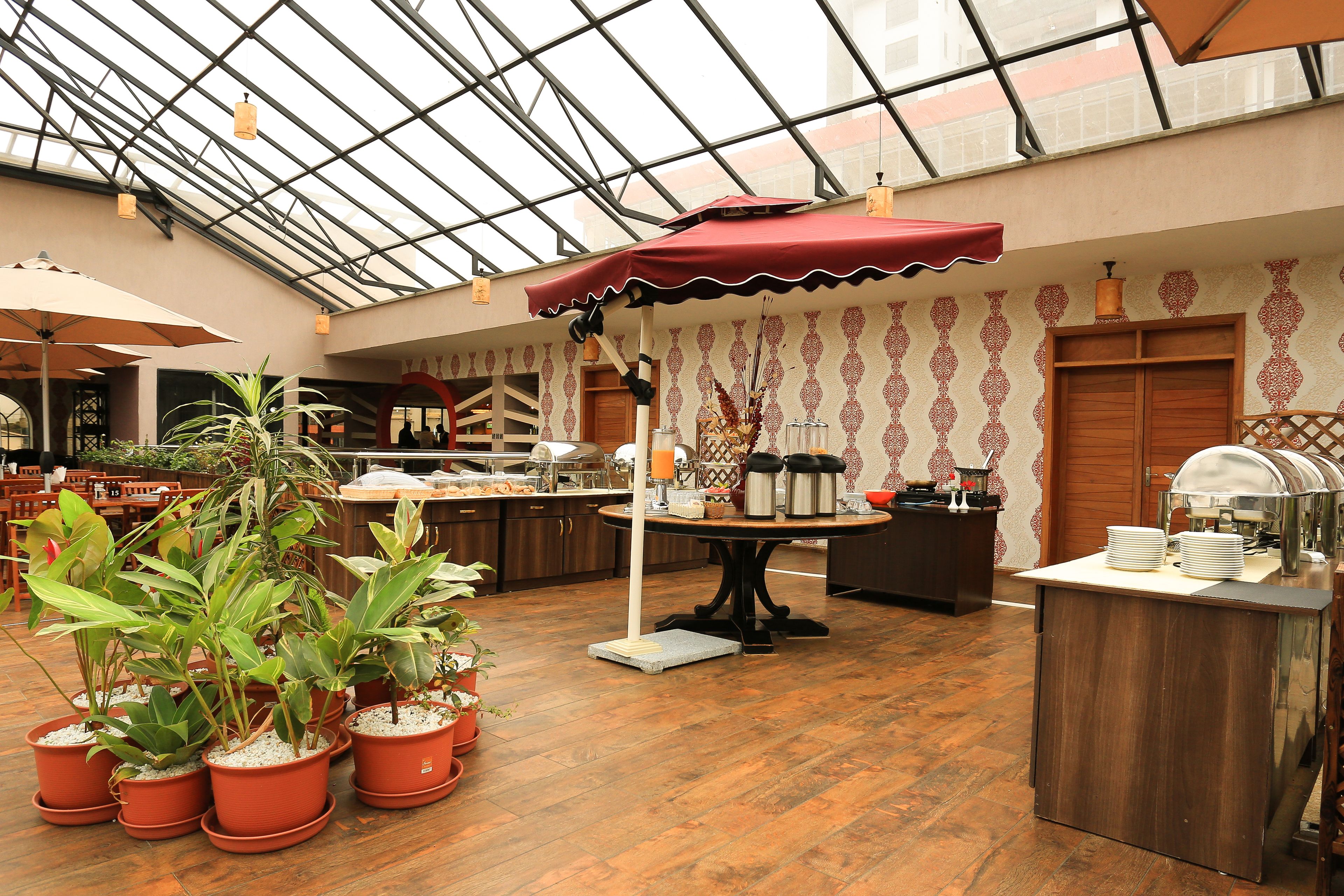 Un vaste espace intérieur doté d’un haut plafond et d’un toit en verre, avec une table centrale surmontée d’un parasol rouge, entourée de plantes en pot et d’un buffet.