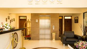 A hotel reception area with a reception desk, a gold stanchion, a red carpet, an elevator, a black leather sofa, and a vase of flowers.