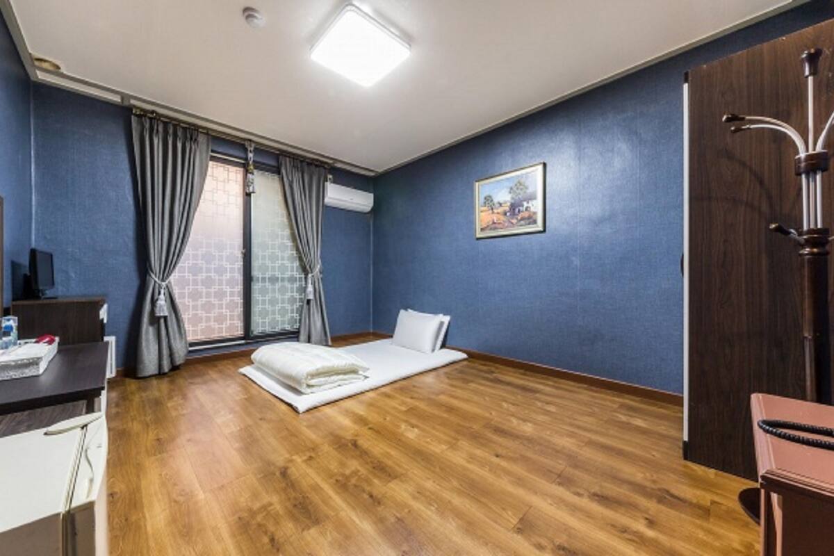 Habitación de hotel con piso de madera, una cama, un escritorio con silla, un televisor y una ventana con cortinas.