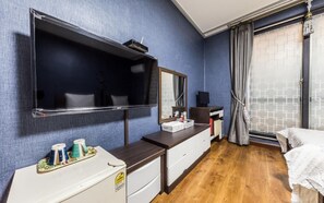 Una camera d'albergo con un grande specchio, una TV a schermo piatto, un piccolo frigorifero, un comodino con articoli da toilette e una finestra vista città.