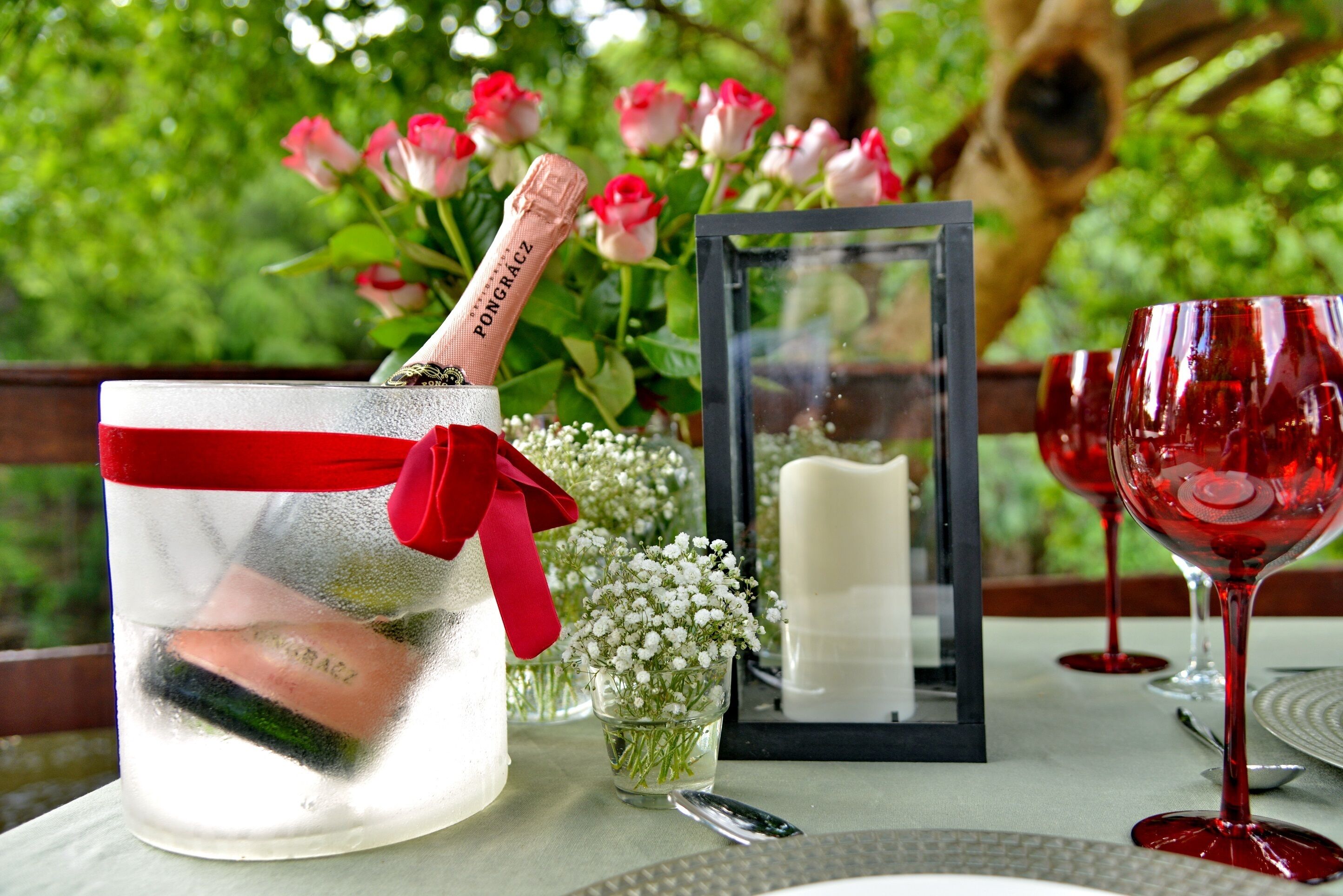 A table set with a candle, a glass of red wine, a bottle of champagne, and a vase with flowers.