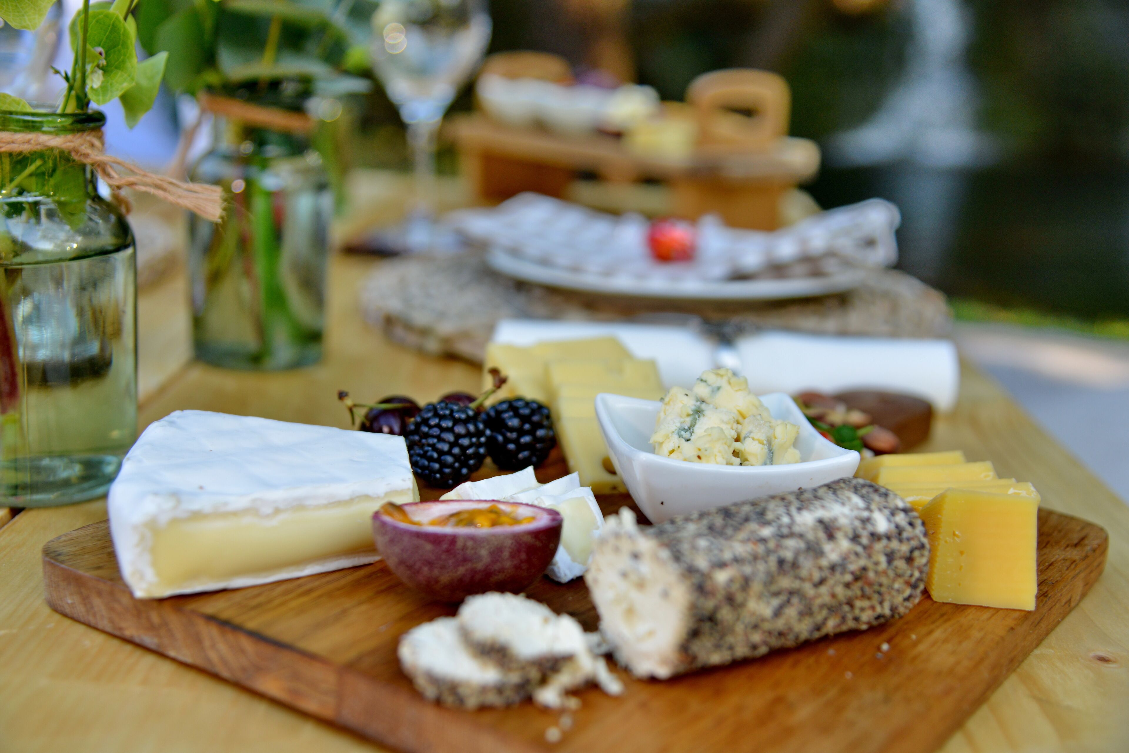 A variety of cheeses and fruits on a wooden board, with a glass of wine and jars in the background.
