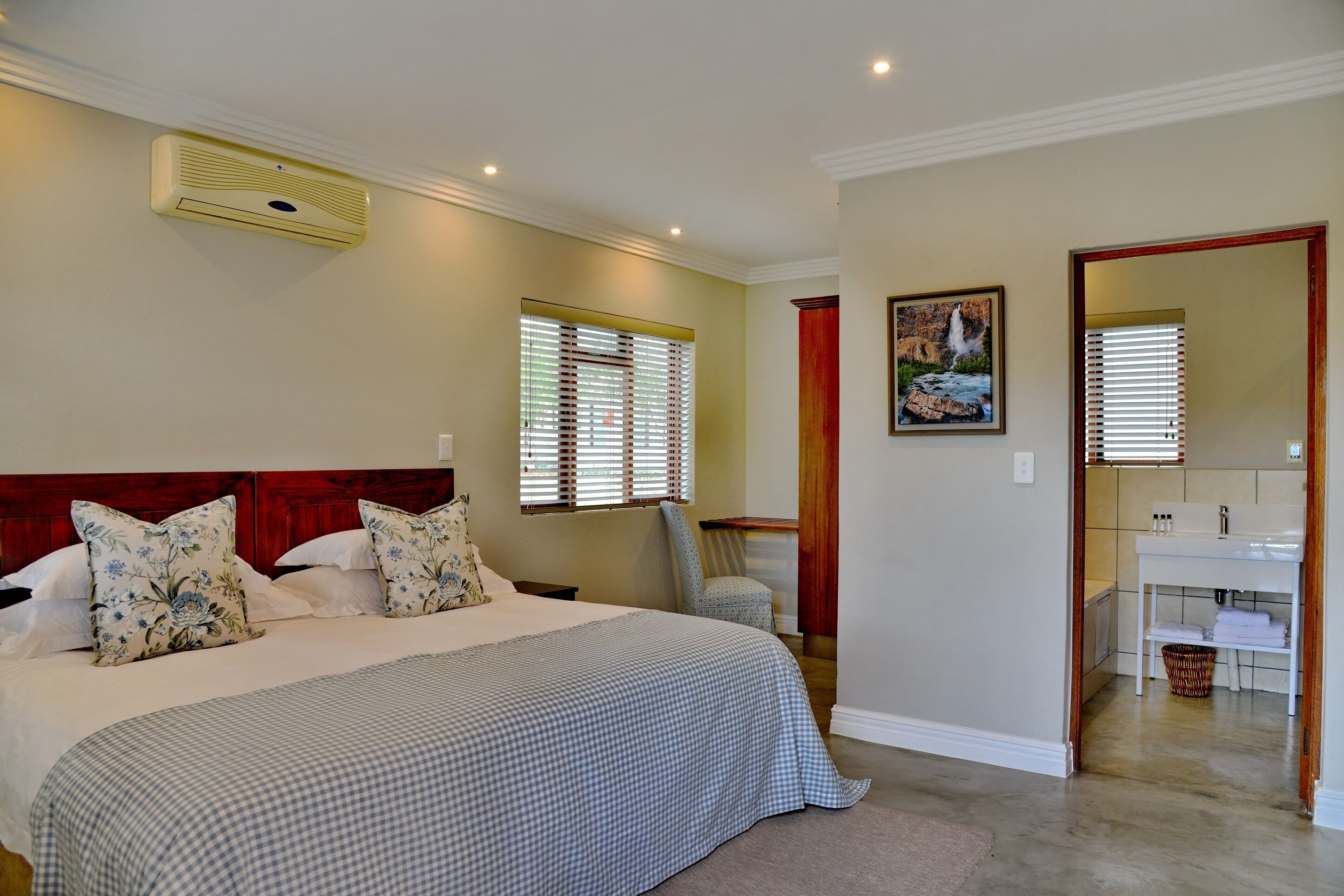 A bedroom with a large bed, a bedside table, a chair, a window with blinds, a painting, and a bathroom visible through an open door.