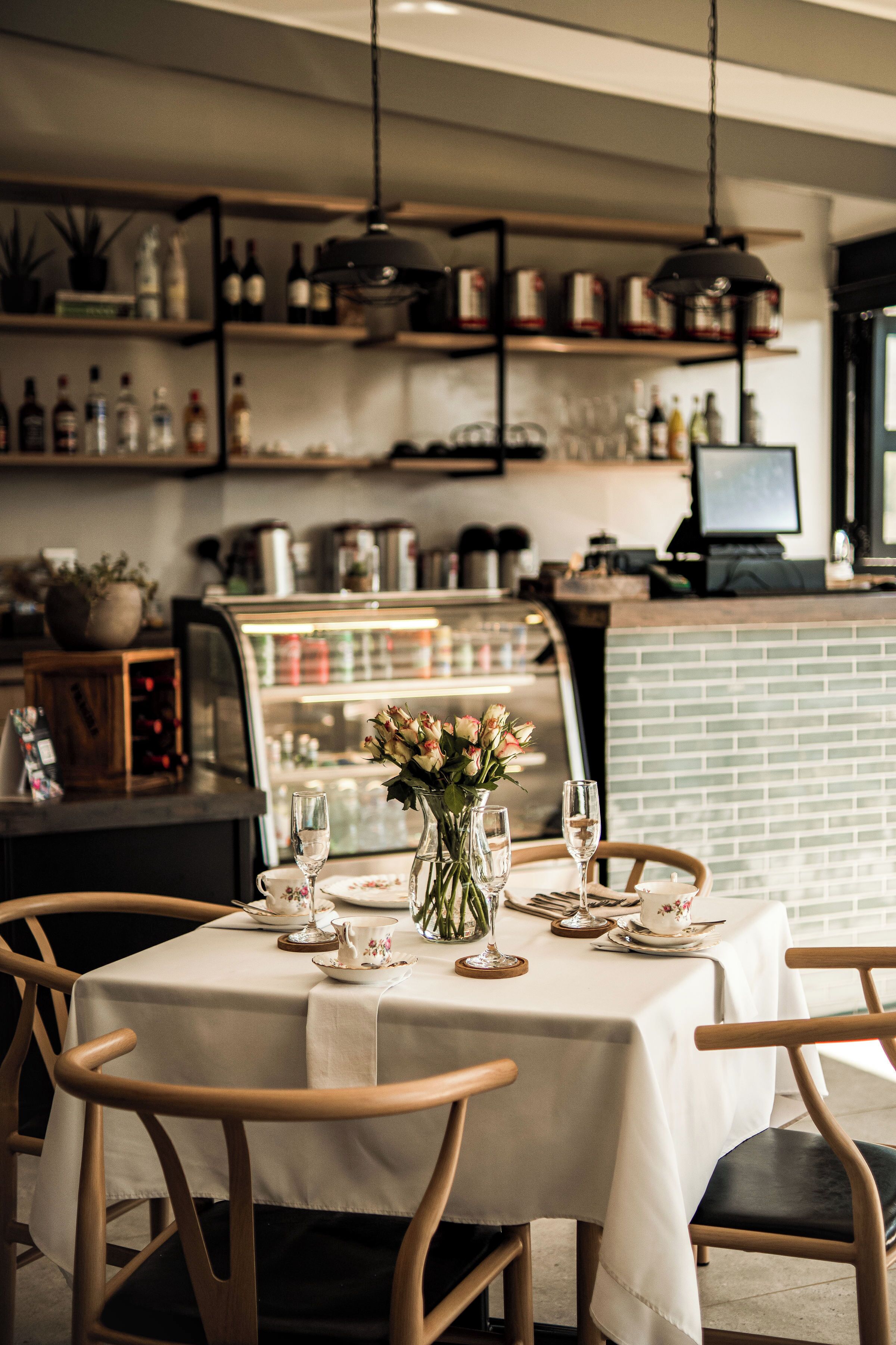 Un interior de restaurante con una mesa lista para dos, un jarrón de flores y estantes llenos de botellas y frascos.