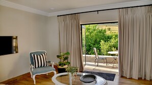 A room with a wooden floor, a white round table, a blue armchair with striped cushions, a potted plant, and a view of greenery through large windows. - Thanda Manzi Country Hotel (Centurion)
