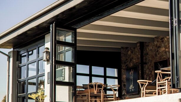 A modern building with a stone exterior, a glass door entrance, outdoor seating area with chairs, and a large potted plant. - Thanda Manzi Country Hotel (Centurion)