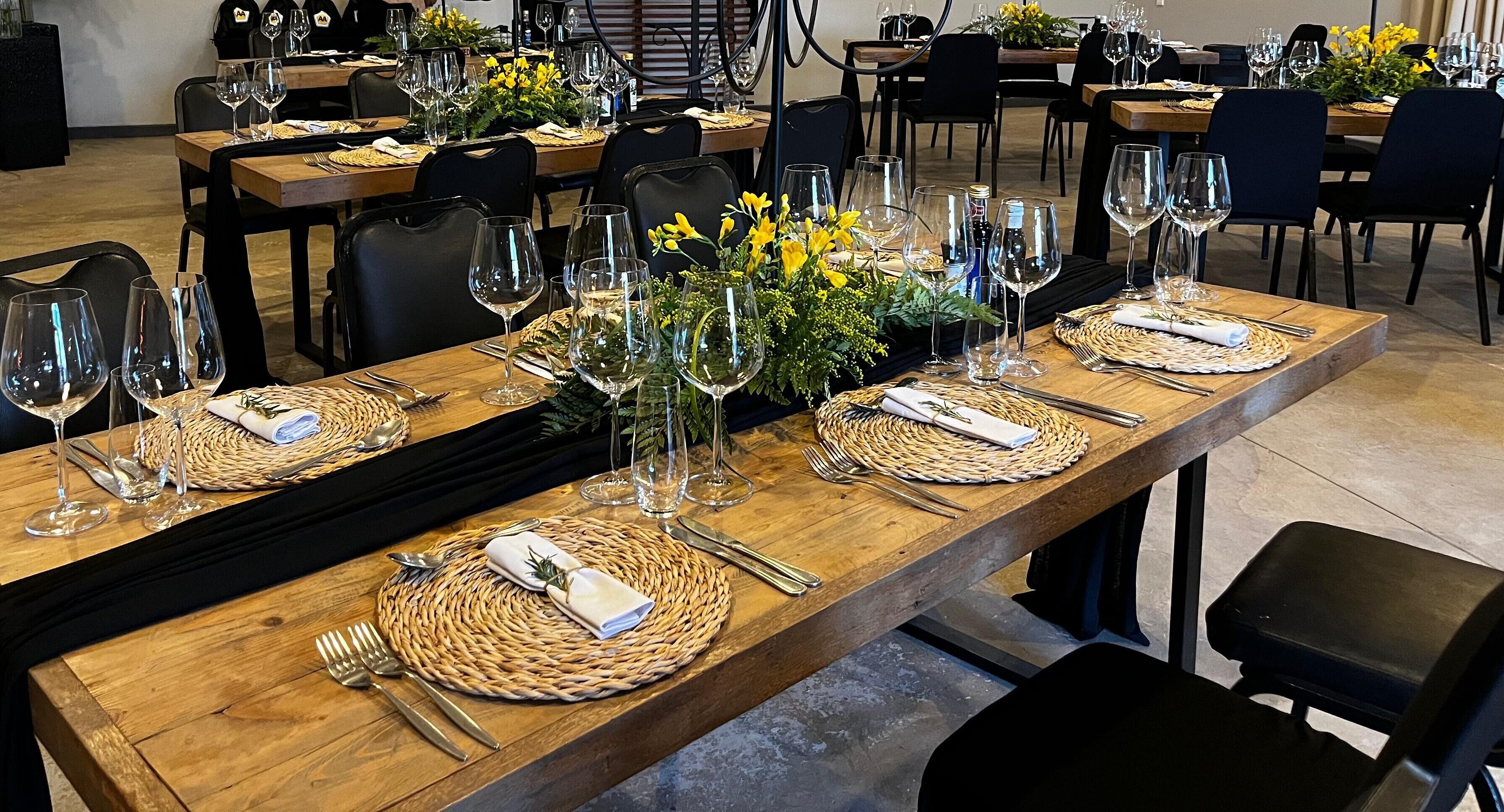 Un restaurante con largas mesas de madera dispuestas para comer, cada una con un posavasos tejido, cubiertos y copas de vino.
