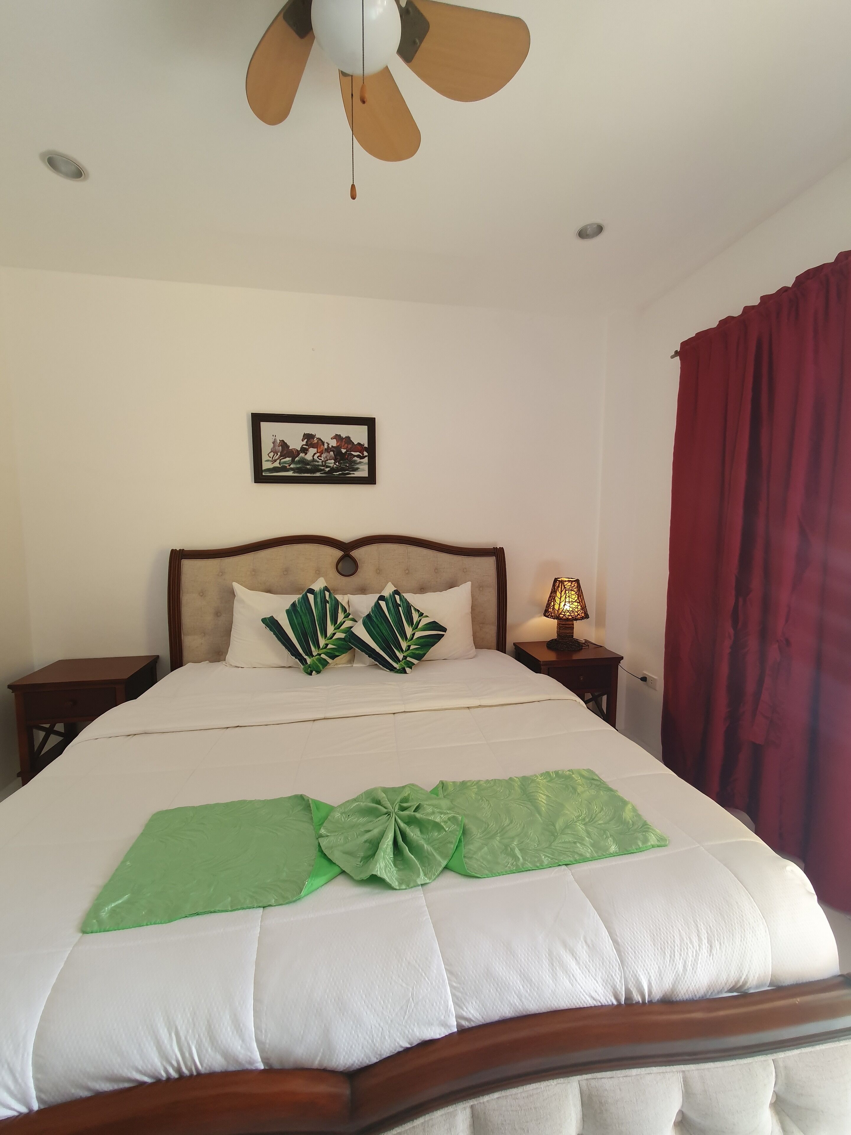 Un lit bien fait, agrémenté d’un coussin décoratif vert, un ventilateur de plafond, un tableau accroché au mur et un rideau rouge.