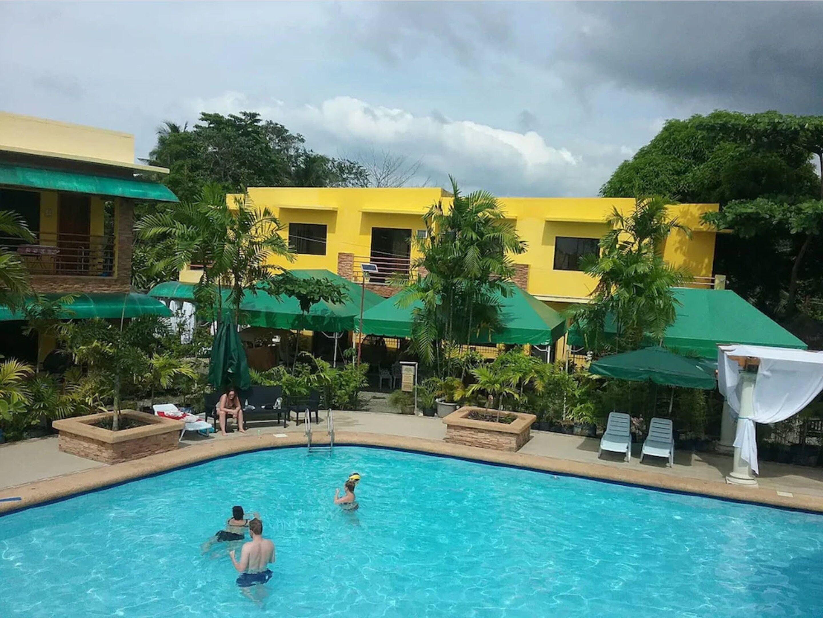 A swimming pool with people in the water, surrounded by green umbrellas and palm trees.