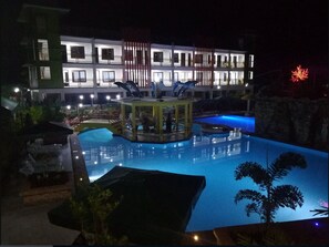 A nighttime swimming pool with a dolphin sculpture, surrounded by a building with multiple balconies and a well-lit facade. - Moalboal Tropics (Moalboal)